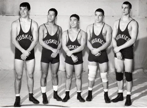 Black-and-white team photo of five young wrestlers standing in a row on a mat, wearing dark singlets lettered "Huskies," knee pads and wrestling shoes.
