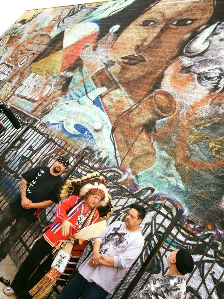 A man in a feathered headdress and red ribbon shirt, holding an eagle feather fan, stands with three men before a large, weathered mural of Indigenous faces on a brick wall.