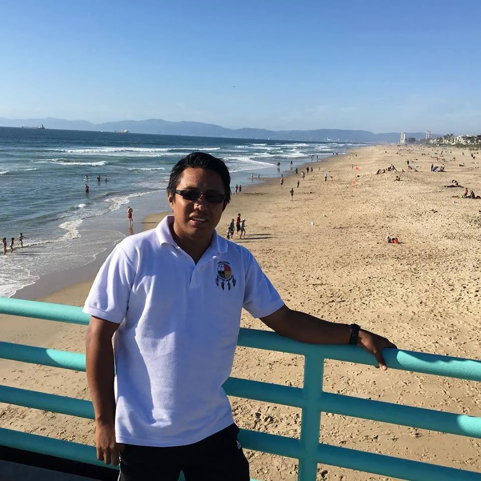 A man in sunglasses and a white polo shirt bearing a Four Worlds medicine wheel emblem leans on a teal railing above a wide sandy beach and ocean.
