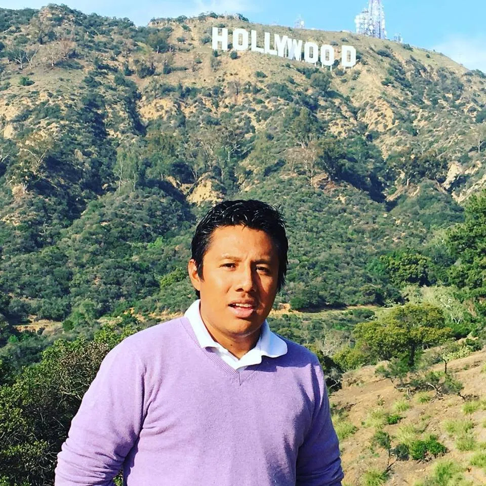 A man in a lavender sweater over a white collared shirt stands before green hills topped by the Hollywood sign.