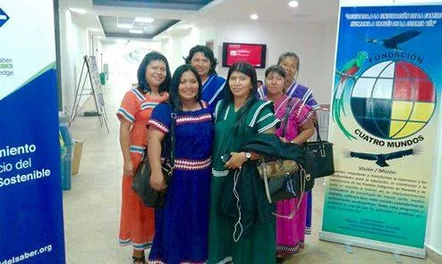 Six Indigenous women in traditional colorful dresses stand together indoors beside a Fundación Cuatro Mundos banner showing a four-color circle and an eagle.