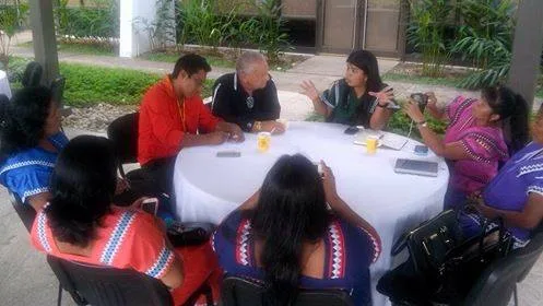 A group seated around a white table outdoors in conversation, several women wearing traditional Ngäbe dresses, one taking photos with a phone.