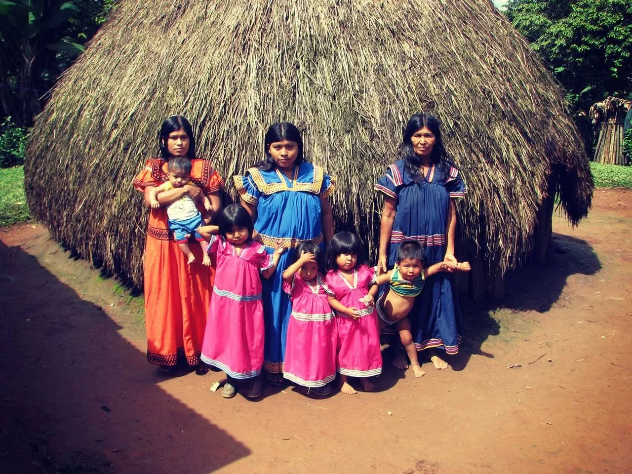 Three women and five small children in traditional bright orange, blue and pink dresses stand before a large thatched round house on red earth.