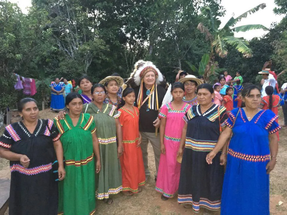 Phil Lane Jr. in a feathered headdress stands outdoors among a group of Ngobe women and girls in brightly colored traditional dresses with zigzag trim.