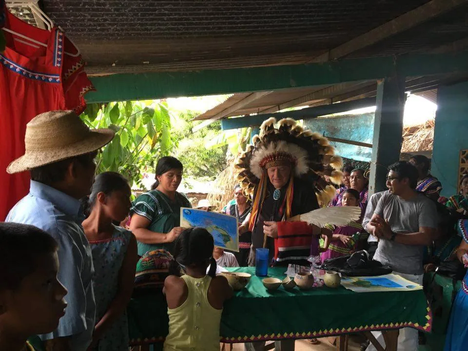 Under a tin roof, Phil Lane Jr. in a feathered headdress stands at a green-covered table as a woman holds a map and community members gather around.