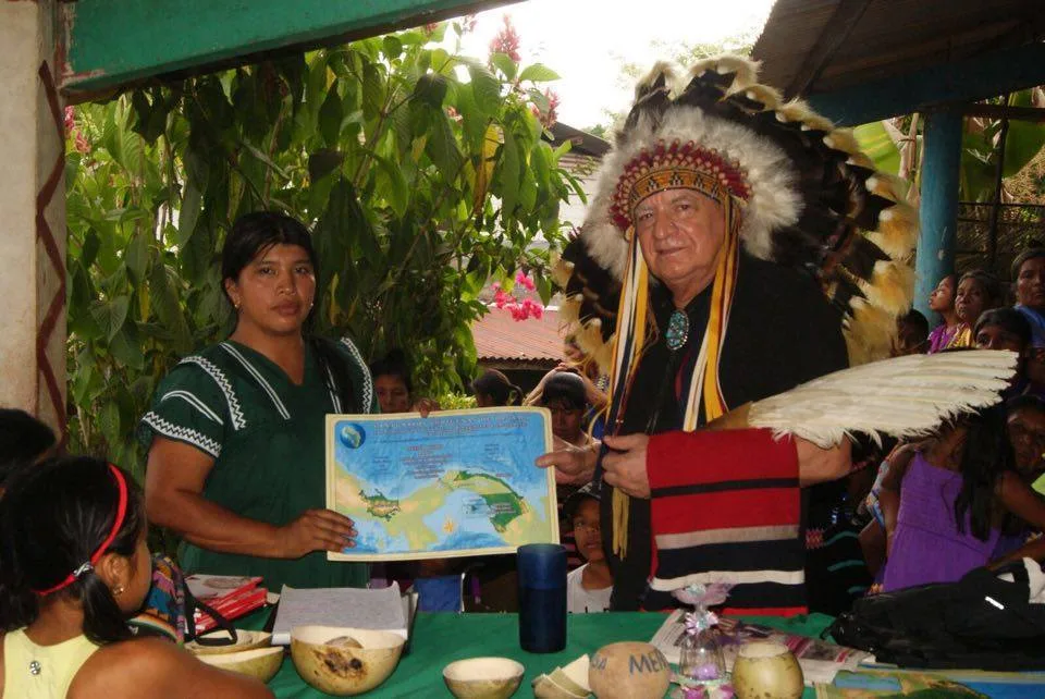 A woman in a green dress with white trim holds a certificate bearing a map of Panama beside an elder in a feathered headdress holding a white feather fan, with a crowd behind them.