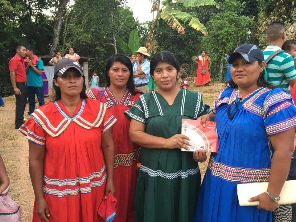 Four Indigenous women in brightly colored traditional dresses with zigzag trim stand outdoors among trees and other people; one holds a copy of El Árbol Sagrado.