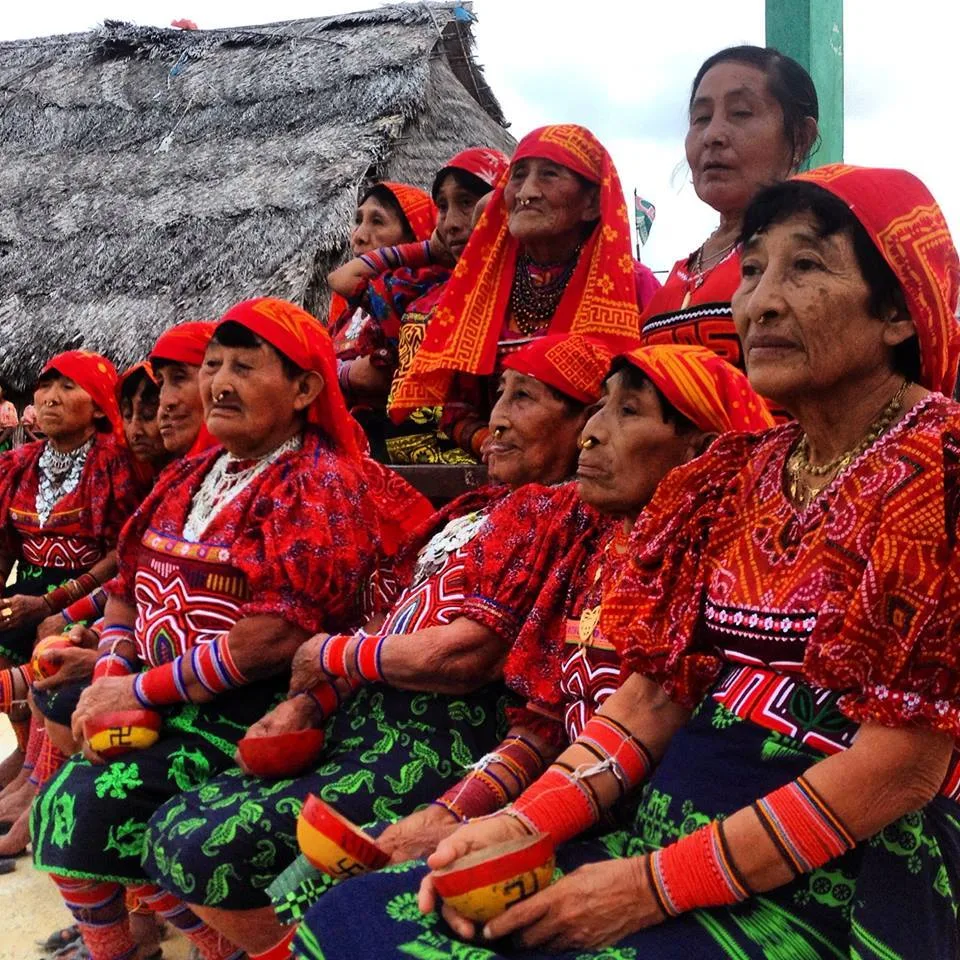 A group of Guna women elders in red headscarves, beaded arm wraps and mola blouses sit together holding painted gourd bowls near a thatched hut.