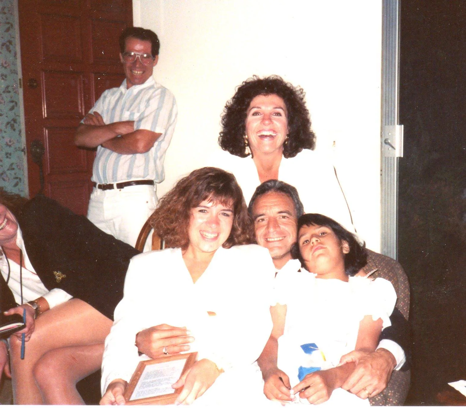 A joyful family gathering: a man seated with two girls in white on his lap, a laughing woman behind them, and others nearby.