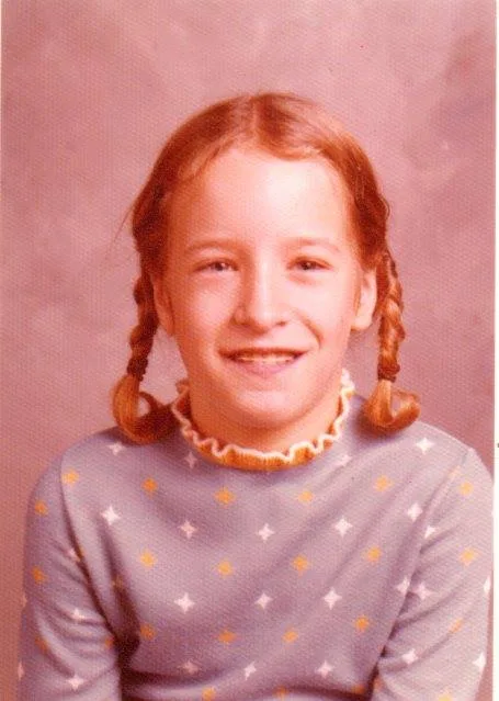 School portrait of a smiling young girl with red braided pigtails wearing a blue patterned top with a ruffled collar.