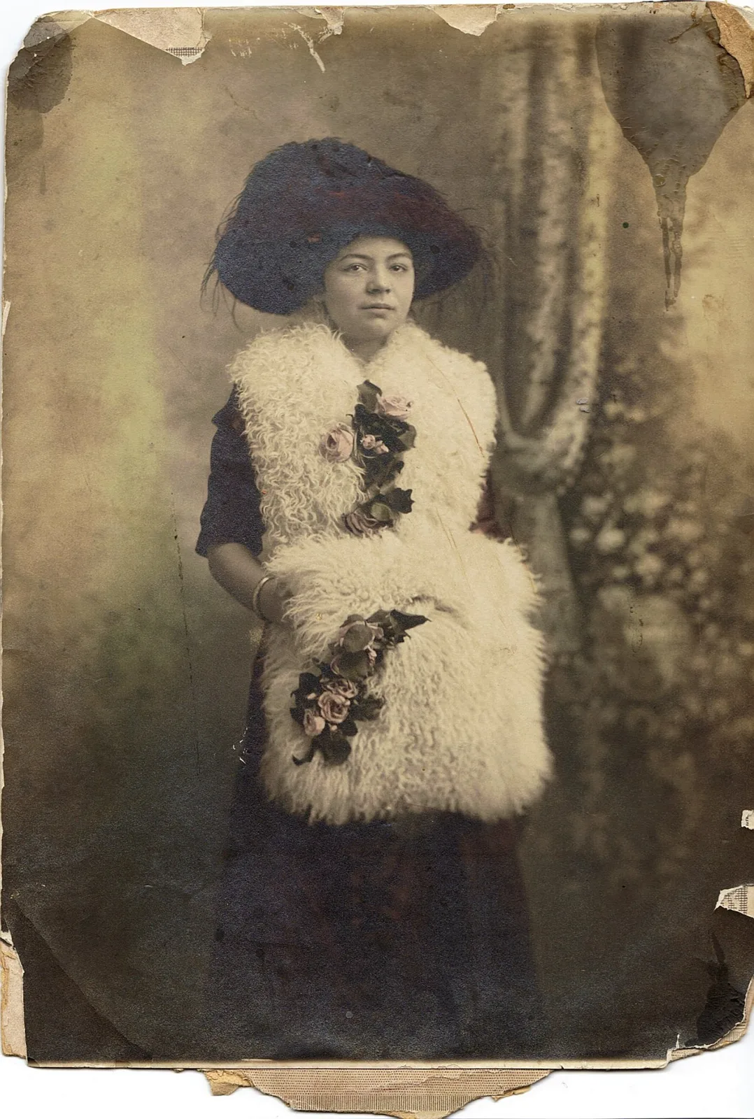 Faded, worn antique portrait of a young woman in a large feathered hat, white fur stole and muff decorated with roses.
