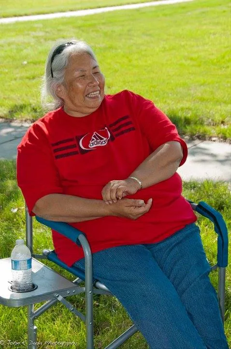 A smiling gray-haired woman in a red T-shirt and jeans sits in a folding chair on a grassy lawn.