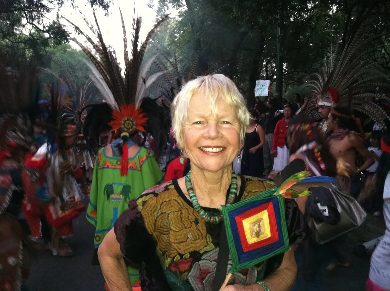 A smiling white-haired woman holding a woven god's eye stands among Aztec dancers in tall feathered headdresses.