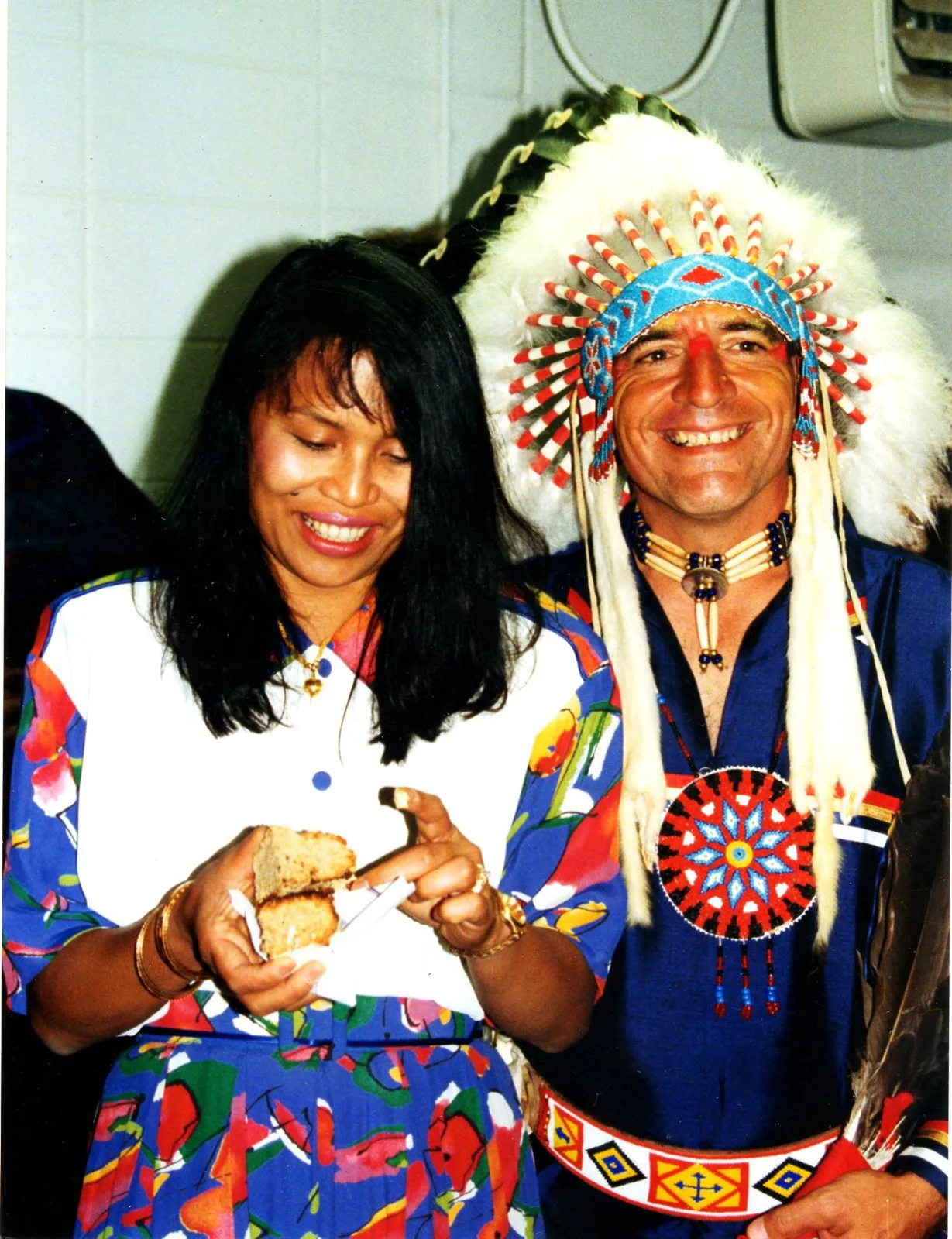A smiling woman holding a piece of bread stands beside a smiling man in a feathered headdress, beaded choker and beaded medallion.