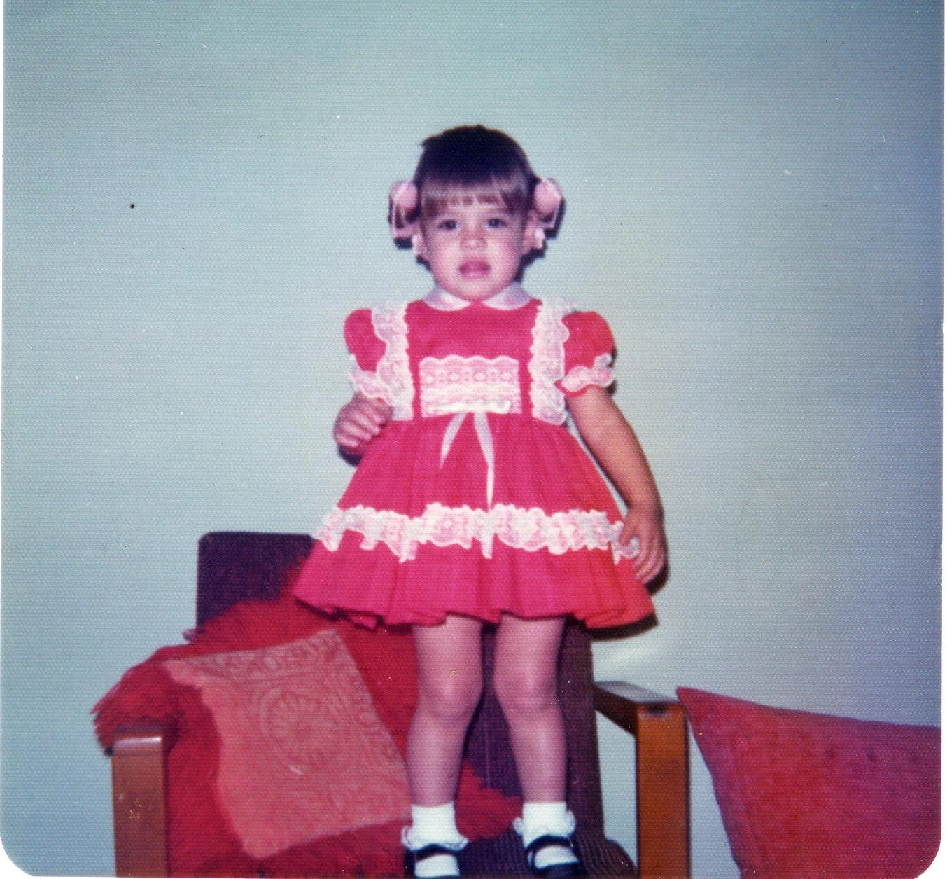 Vintage photo of a toddler girl with pink hair bows in a red dress with white lace trim, standing on a chair.