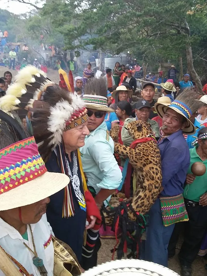 Phil Lane Jr., in a feathered headdress, smiles amid a crowd of Ngäbe Buglé people in colorful hats; a man blows a whistle beside a jaguar pelt held aloft outdoors.