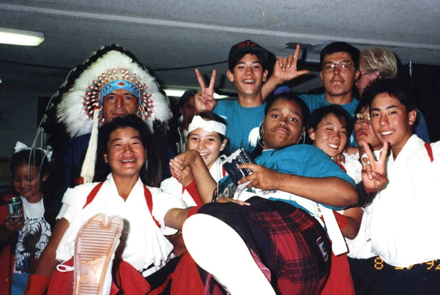 An elder in a large white feather headdress poses with a joyful group of young people, some in white and red uniforms, laughing and making peace signs; dated 1992.