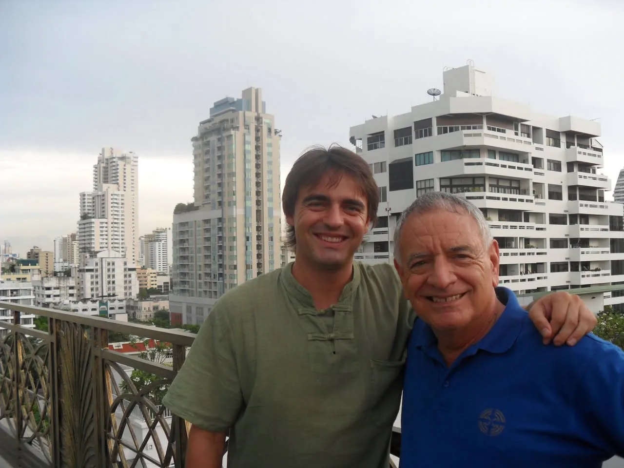 Two smiling men stand arm in arm on a rooftop terrace with high-rise apartment buildings behind them; one wears a green shirt, the older man a blue polo.