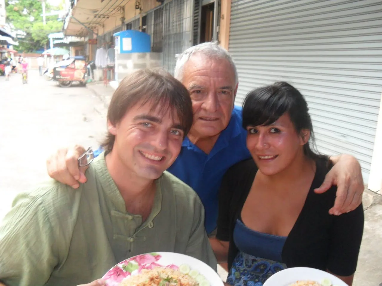 A young man in a green shirt, an older man in a blue polo and a young woman with dark hair smile together at a street-side table with plates of fried rice.