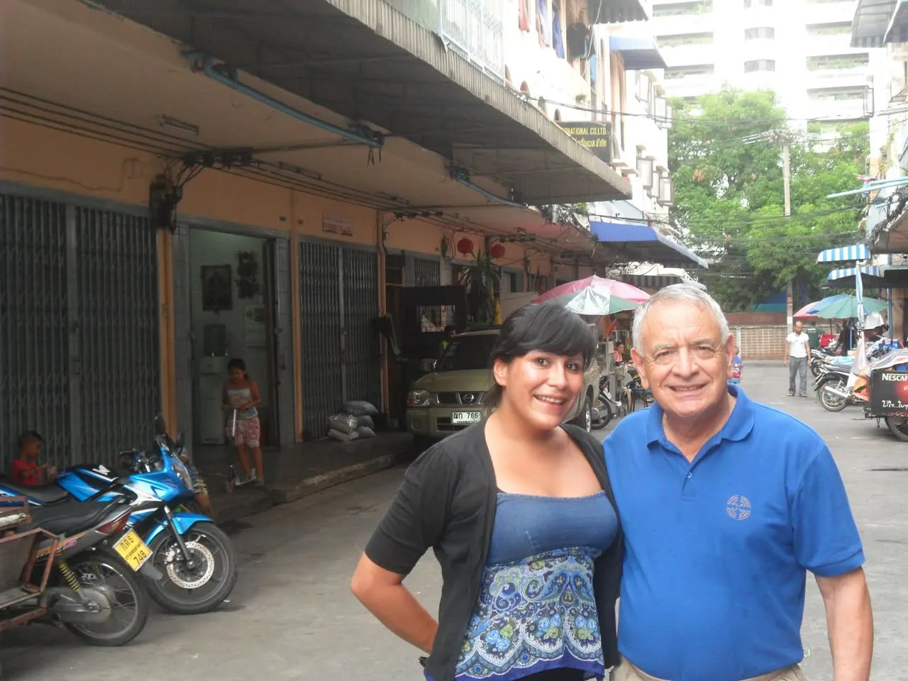 A young woman in a black cardigan and blue patterned top stands beside an older man in a blue polo on a narrow street lined with shops and parked motorbikes.
