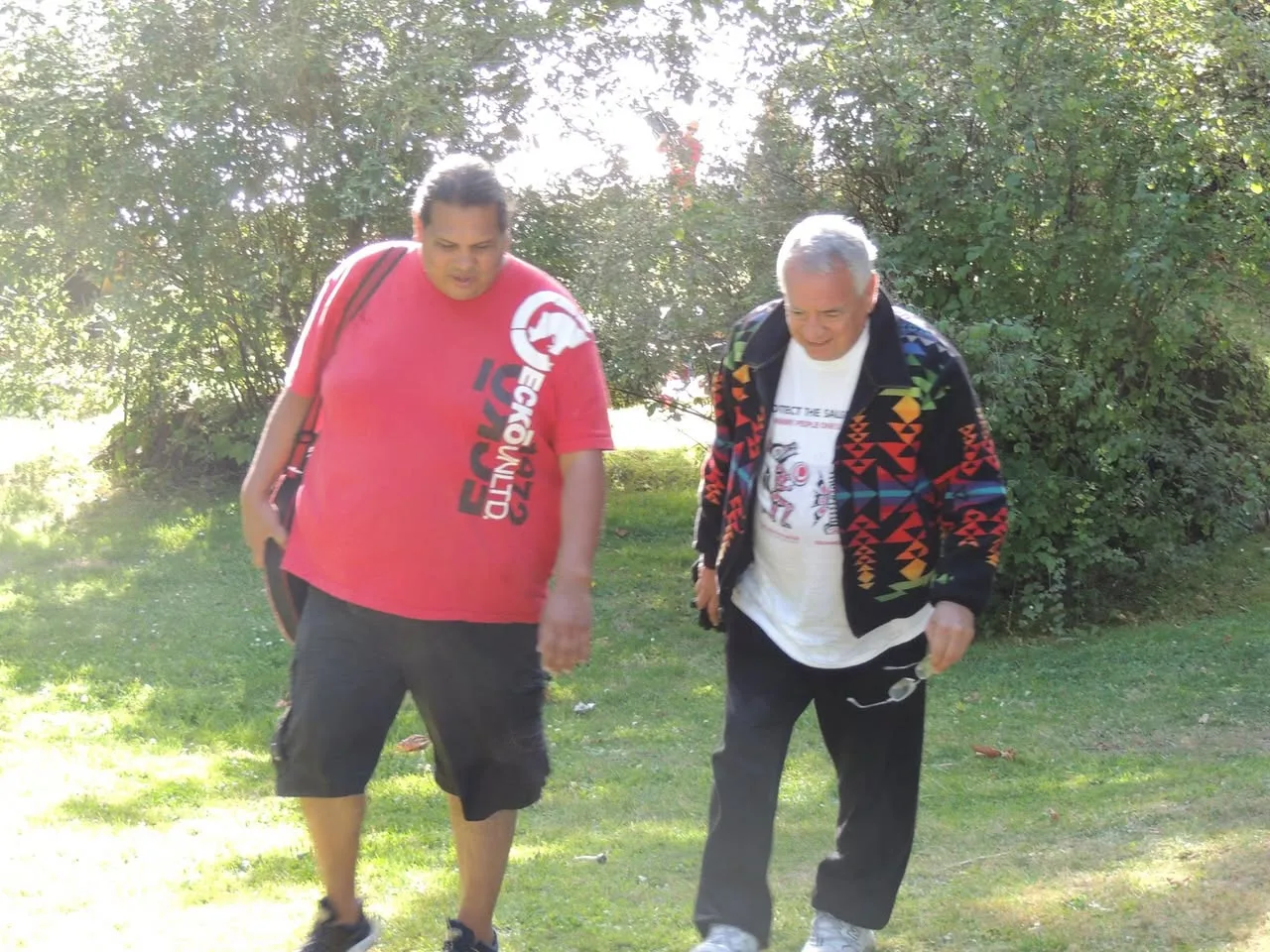 Two men walk across a sunlit lawn beneath trees, one in a red T-shirt carrying a drum case, the other a white-haired elder in a colorful geometric-patterned jacket.