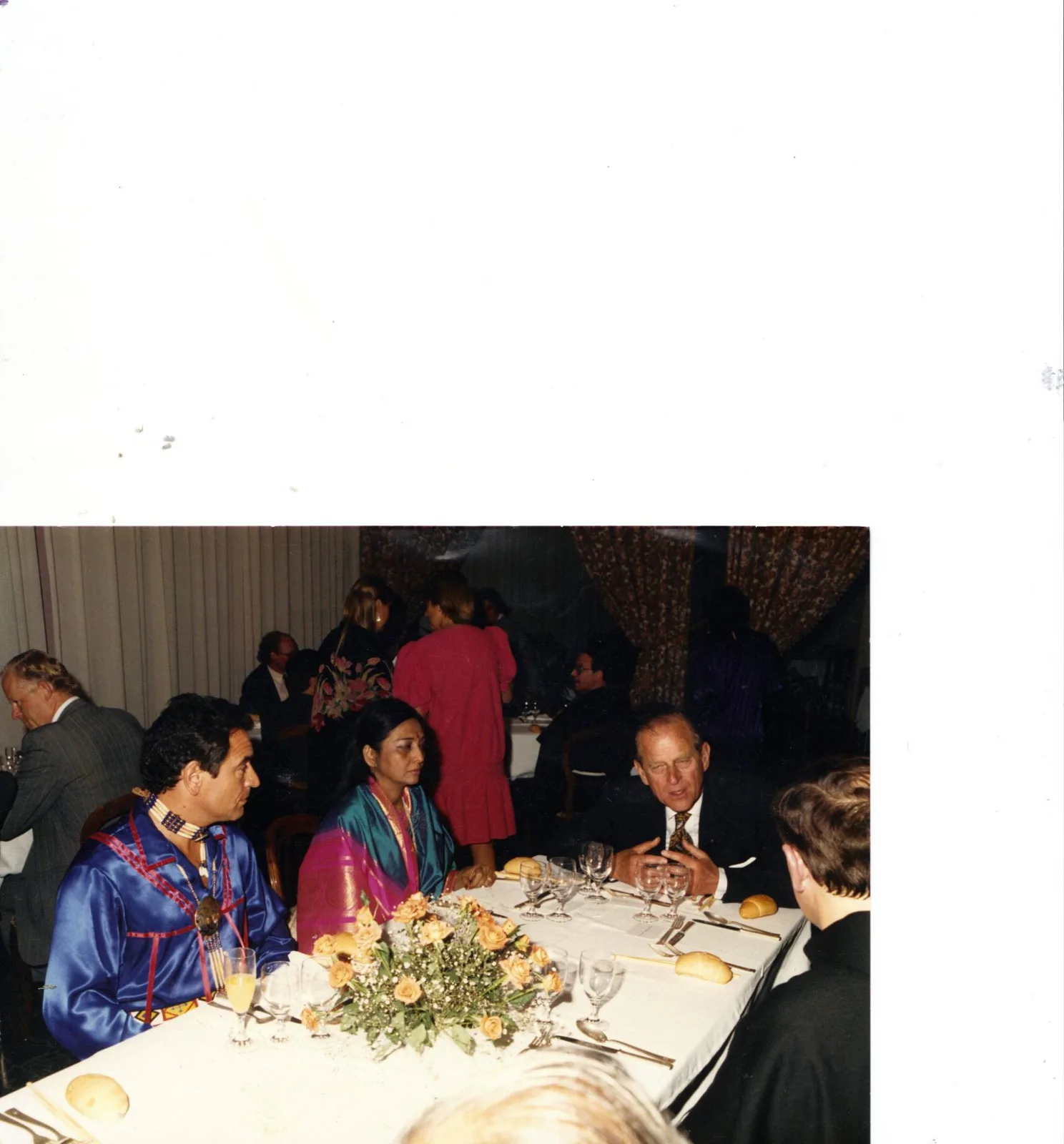At a banquet table with roses, a man in a blue ribbon shirt and beaded choker sits beside a woman in a teal and pink sari, listening to an older man in a dark suit speaking.