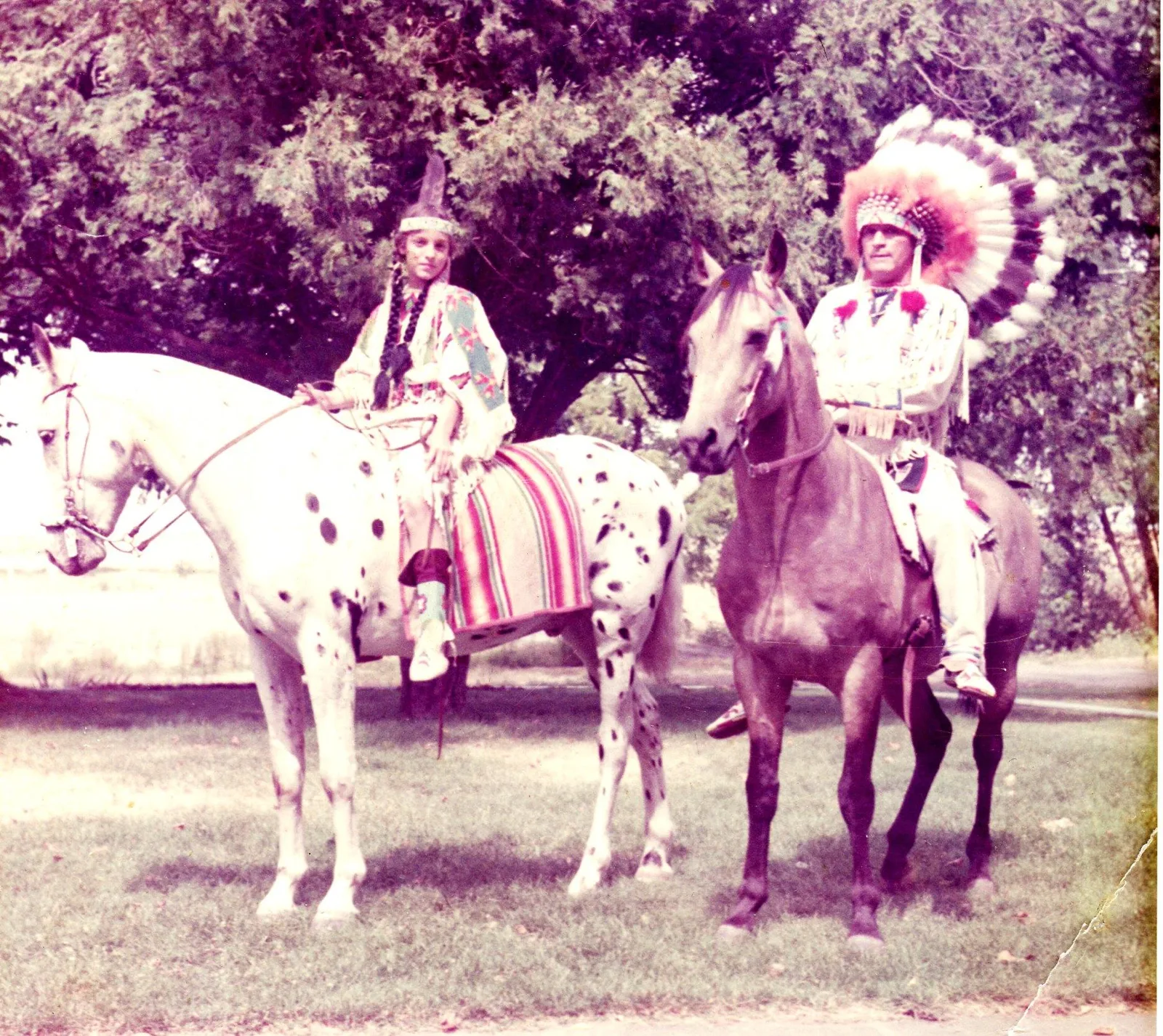 Faded color photo of a child in a beaded dress and feather headband on a spotted horse beside a man in a feather war bonnet on a gray horse.