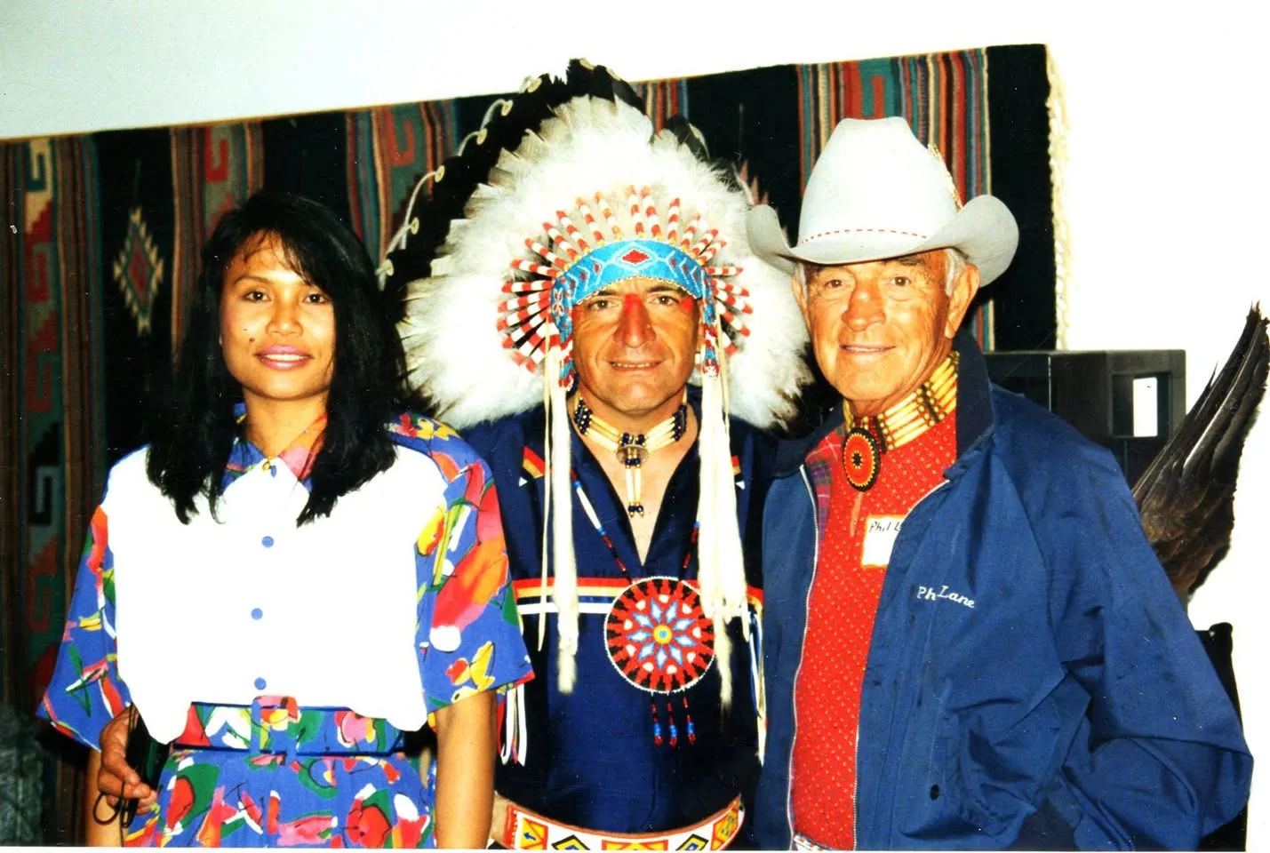 A woman in a floral dress, a man in a feather headdress and beaded regalia, and Phil Lane Sr. in a cowboy hat and jacket marked Ph Lane.