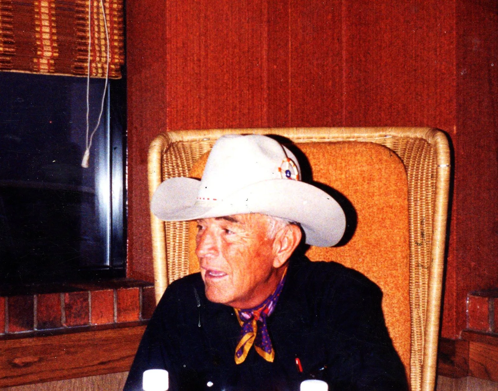 An elder in a white cowboy hat and patterned neckerchief sits in a wicker chair, smiling and looking to the side.