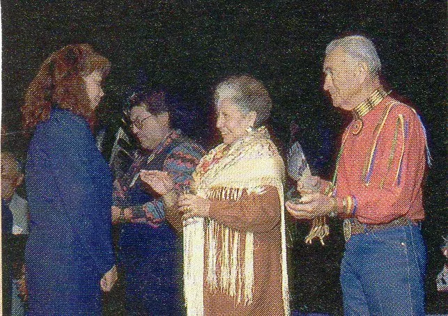 Four people at an evening event: a woman in a fringed shawl and a man in a ribbon shirt reach toward a red-haired woman.