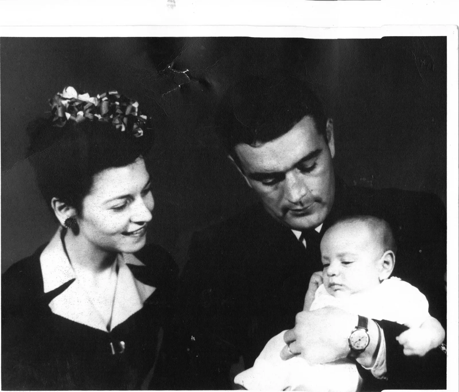 Vintage black and white portrait of a smiling woman beside a man in a suit who looks down at a baby in his arms.