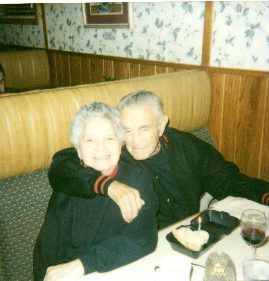 An elderly couple embrace and smile in a restaurant booth, with a candle-topped dessert and a glass of wine on the table.
