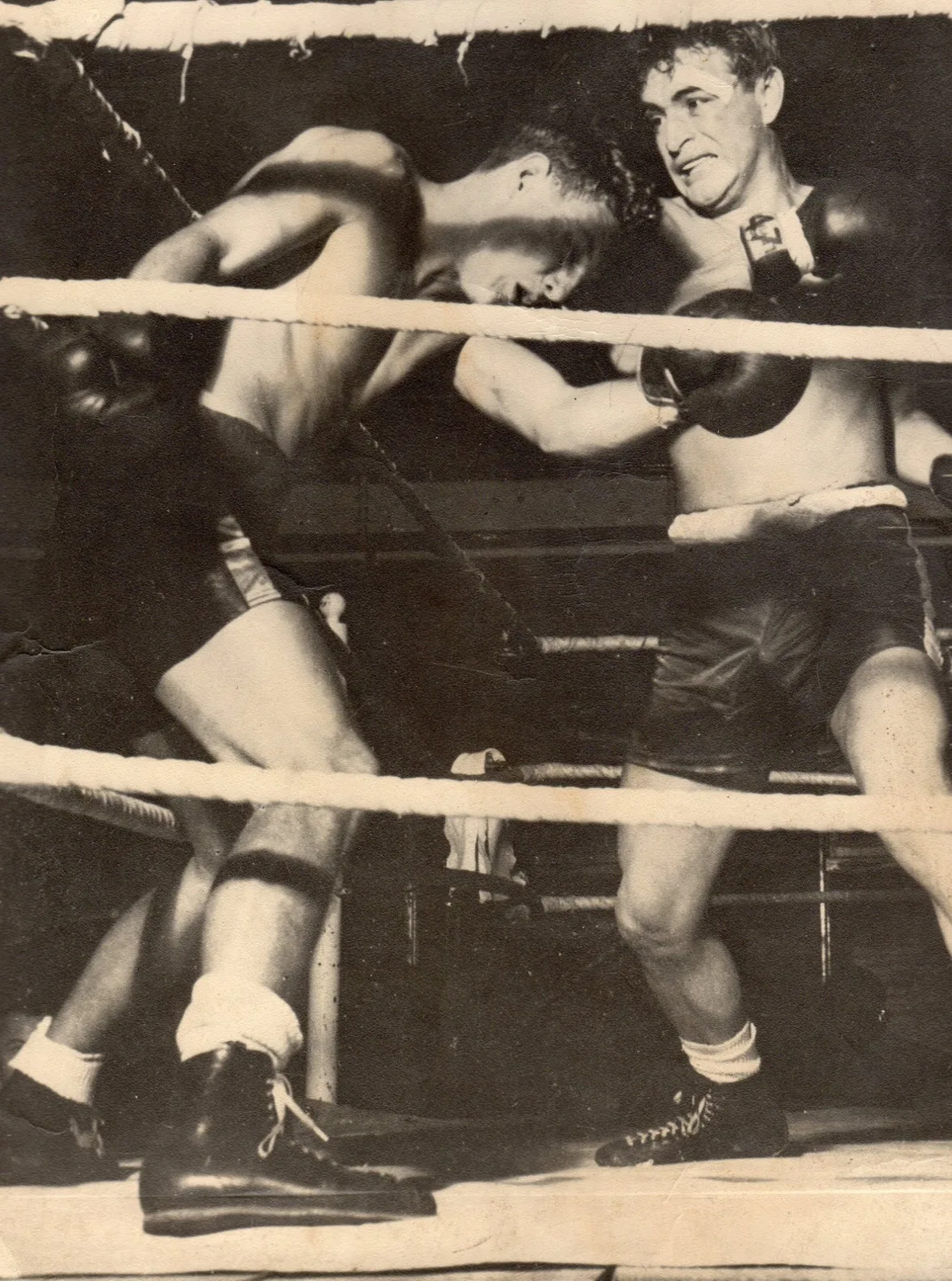 Vintage black-and-white photograph of two boxers fighting in a ring, one leaning forward against the ropes as the other throws a punch.