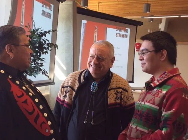 Three men stand talking and smiling in a bright hall: one in a black jacket with red designs, a white-haired elder with a turquoise bolo tie, and a young man in a patterned red jacket.