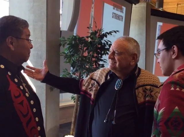 The white-haired elder in a patterned cardigan and turquoise bolo tie gestures warmly toward the man in the black and red jacket as the young man listens, with a "Spiritual Strength" banner behind them.