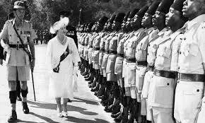 Black-and-white photograph of a woman in a white dress and hat inspecting a long line of uniformed African soldiers, escorted by a colonial officer.