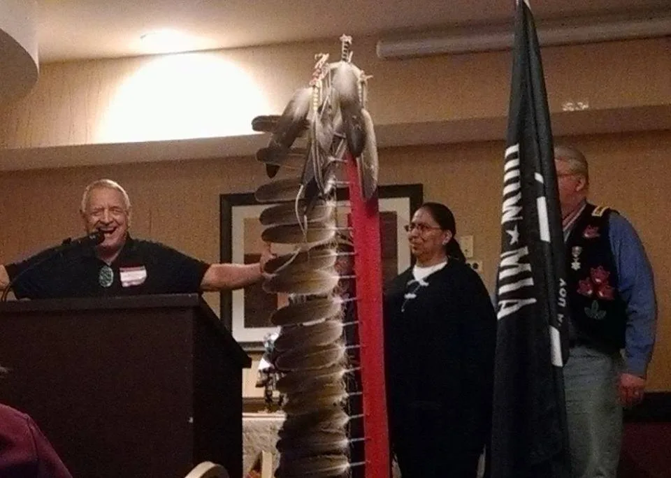 A smiling white-haired man speaks at a podium beside a tall eagle feather staff, with a woman and a man in a beaded vest standing near a POW-MIA flag.