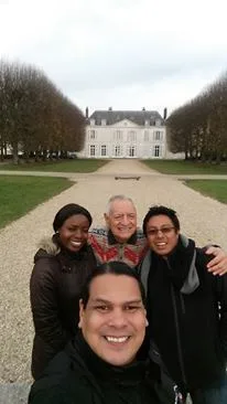 Four smiling people pose for a selfie on a gravel path lined with bare trees, with a large white château behind them on a cloudy day.