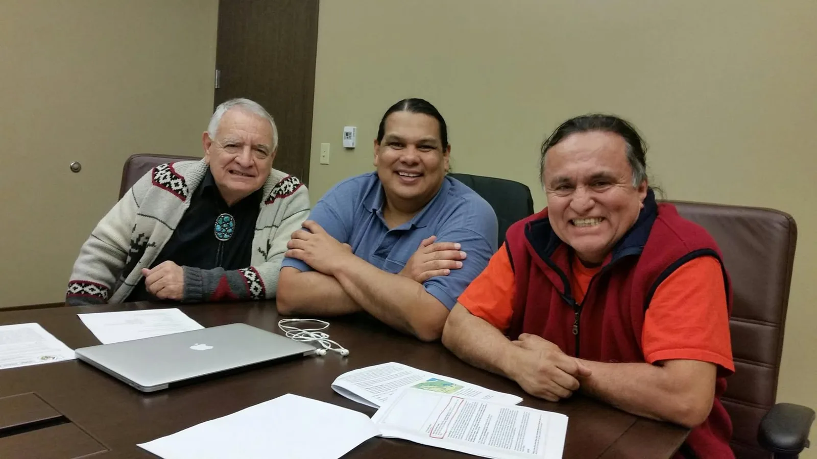Three smiling Indigenous men seated at a conference table with a laptop, earphones and printed documents in front of them, in a meeting room with beige walls.