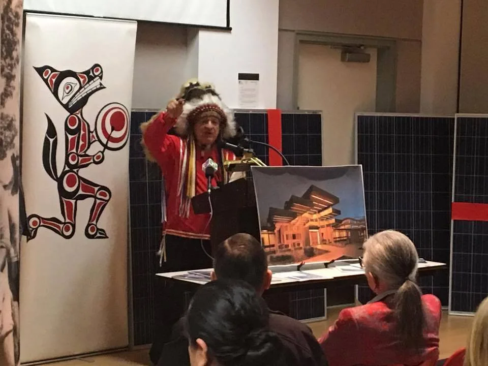 An elder in a feathered headdress and red ribbon shirt raises his fist while speaking at a podium, beside a Coast Salish-style wolf banner, solar panels and a building rendering.