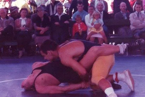 A vintage photo of two wrestlers on a mat, one in a dark singlet and yellow tights pinning his opponent, as spectators of all ages watch from benches behind them.
