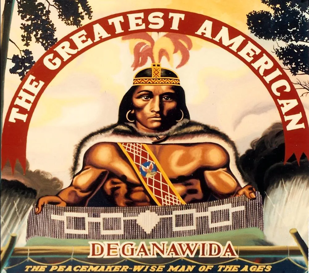 Painting of Deganawida beneath a red banner reading "The Greatest American," wearing a fur cape and holding a wampum belt; text below reads "Deganawida, The Peacemaker, Wise Man of the Ages."