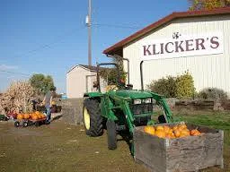 A green tractor sits in front of a white building with a sign reading "Klicker's," beside a wooden crate of orange pumpkins.