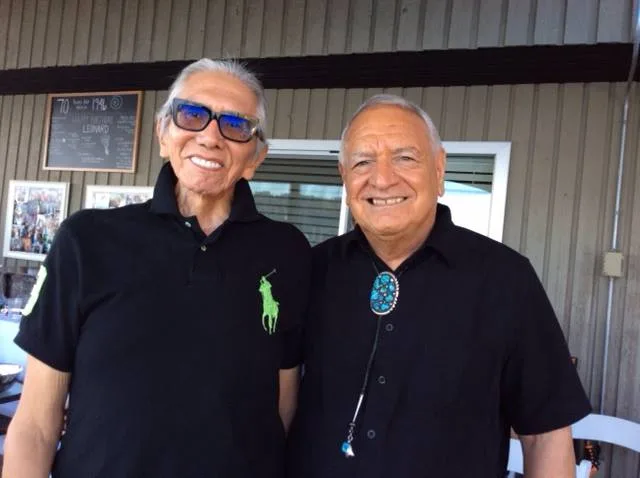 Two smiling Indigenous elders, Leonard George and Phil Lane Jr., in black shirts on an outdoor patio, one in blue sunglasses, the other in a turquoise bolo tie.