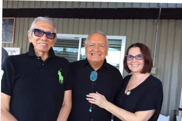 Leonard George and Phil Lane Jr. smiling on a patio beside a woman with short auburn hair and glasses, who rests her hand on the arm of the man in the bolo tie.