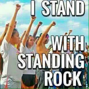 A crowd of people, many young women, raise clenched fists against a blue sky, with bold white text reading "I Stand With Standing Rock."