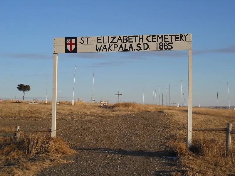 A white wooden archway over a gravel road reads "St. Elizabeth Cemetery, Wakpala, S.D. 1885" with a cross shield, crosses and grave markers on dry prairie beyond.