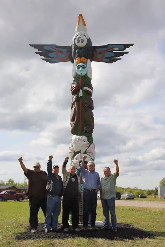 Five men stand with fists raised in front of a tall, brightly painted totem pole topped by an eagle with outstretched wings, on a grassy field under a cloudy sky.