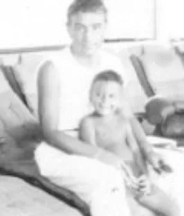 Faded black-and-white photo of a man in a white sleeveless shirt sitting with a smiling young boy on his lap, likely Phil Lane Jr. with his father.