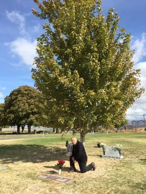 A man in black kneels beside a grave marker with red roses in a sunny cemetery, under a tall leafy tree and blue sky with clouds.